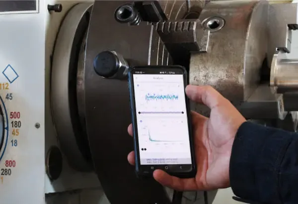 A technician holding a phone with WiSER VIBE® showing a time waveform and a spectrum, in front of a lathe.